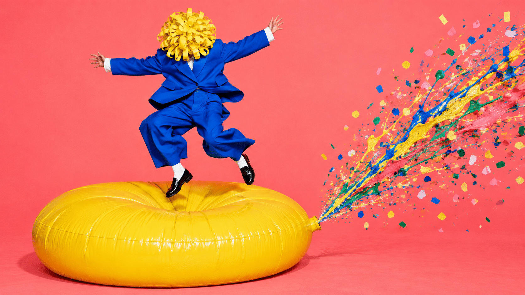 An artist in a blue suit and yellow ribbon hood jumps on a giant yellow whoopee cushion, sending paint and confetti across a coral pink studio.